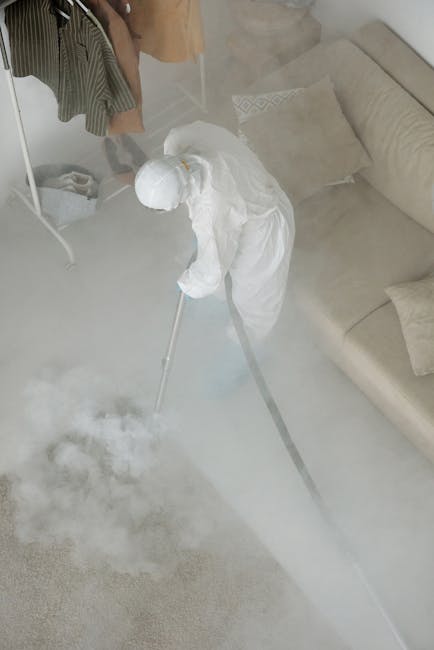A person wearing black gloves is using a yellow handheld steam cleaner on a dark gray fabric sofa in a domestic setting. The sofa has a modern design with wide armrests and a slightly textured surface