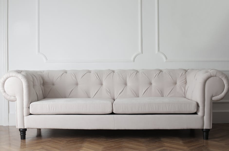 A white upholstered sofa with button-tufted detailing along the backrest and rolled arms, positioned against a plain white wall with decorative molding. The sofa rests on dark wooden legs and is place